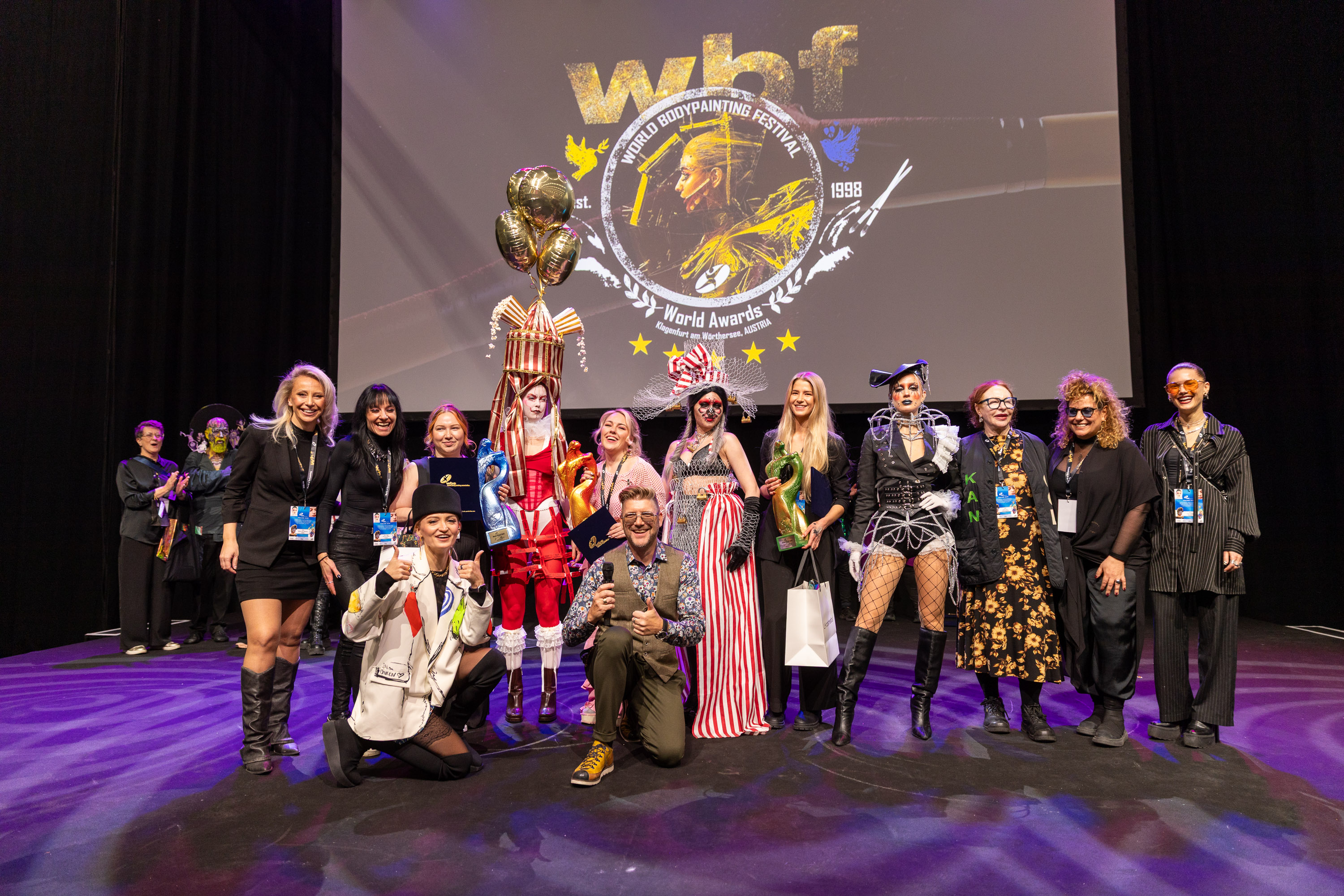 Frau in Leoparden-Bodypainting und großem, mit Früchten geschmücktem Hut posiert am Wasser in der Sonne.