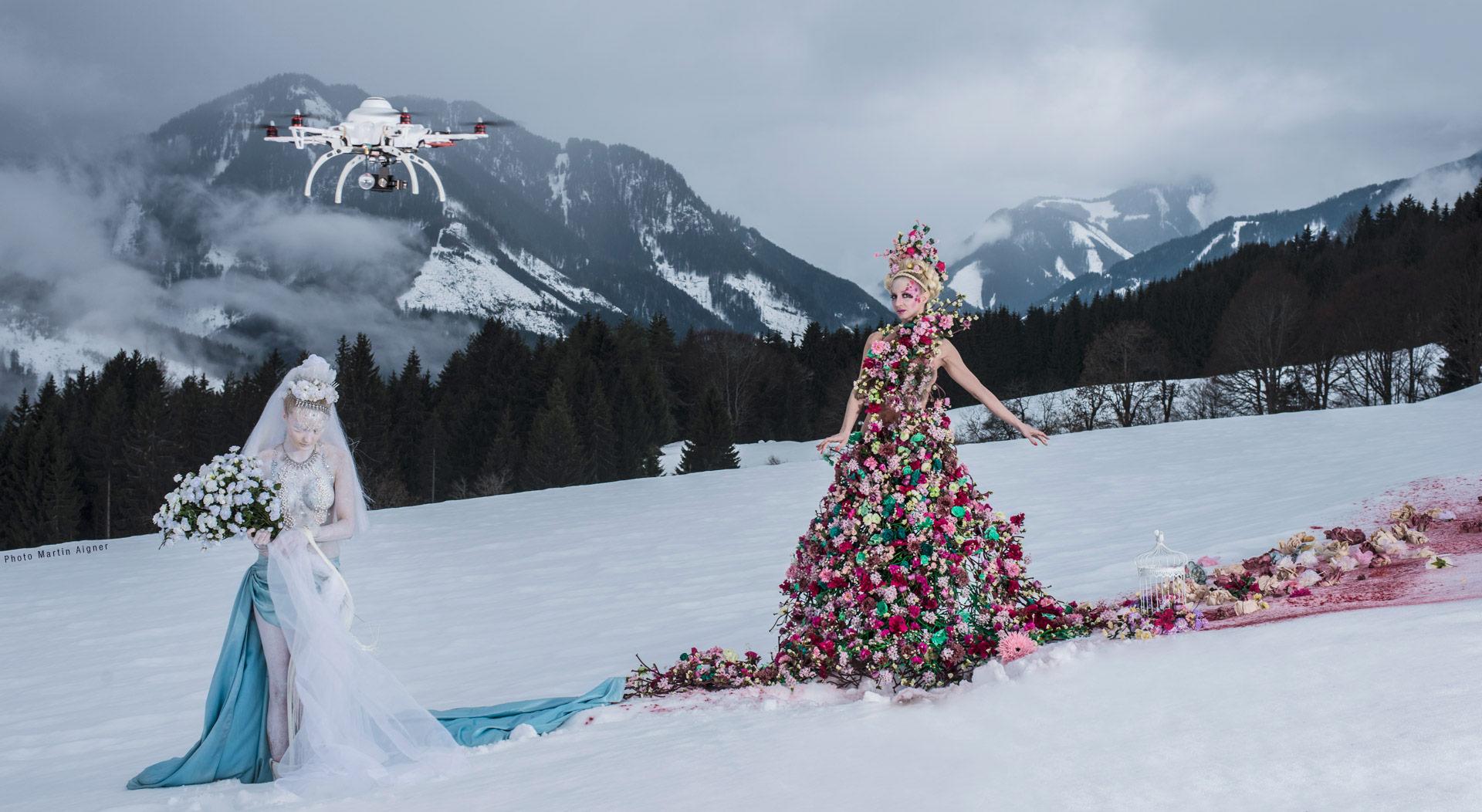 Zwei Figuren in kunstvollen Kostümen stehen in einer verschneiten Berglandschaft: links eine weiß gekleidete Frau mit Blumenstrauß, rechts eine Frau in einem Kleid aus bunten Blumen. Am Himmel schwebt eine Drohne.