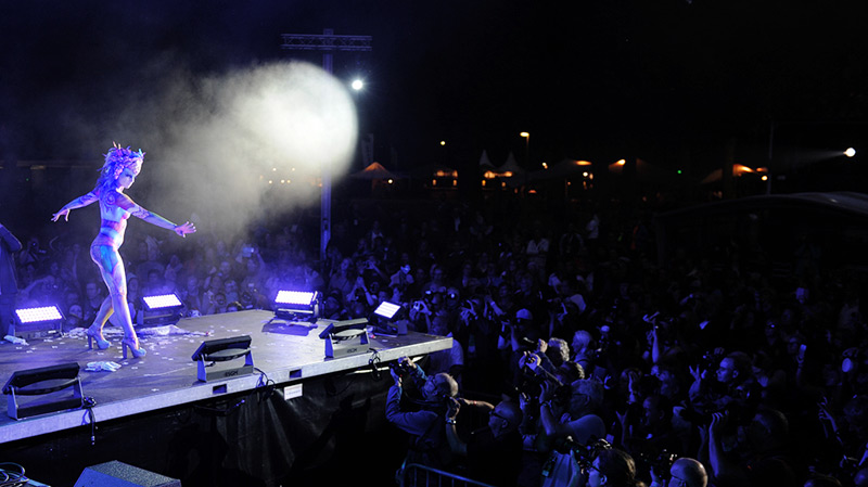 Showauftritt bei Nacht: Eine bemalte Performerin schreitet über eine hell erleuchtete Bühne, während Fotografen und ein großes Publikum die Szene verfolgen.