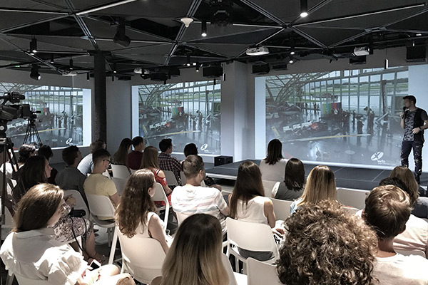 Publikum in einem modernen Konferenzraum verfolgt eine Präsentation. Auf der Leinwand sind Projektionen von Bodypainting-Performances zu sehen, während ein Sprecher auf der Bühne steht.