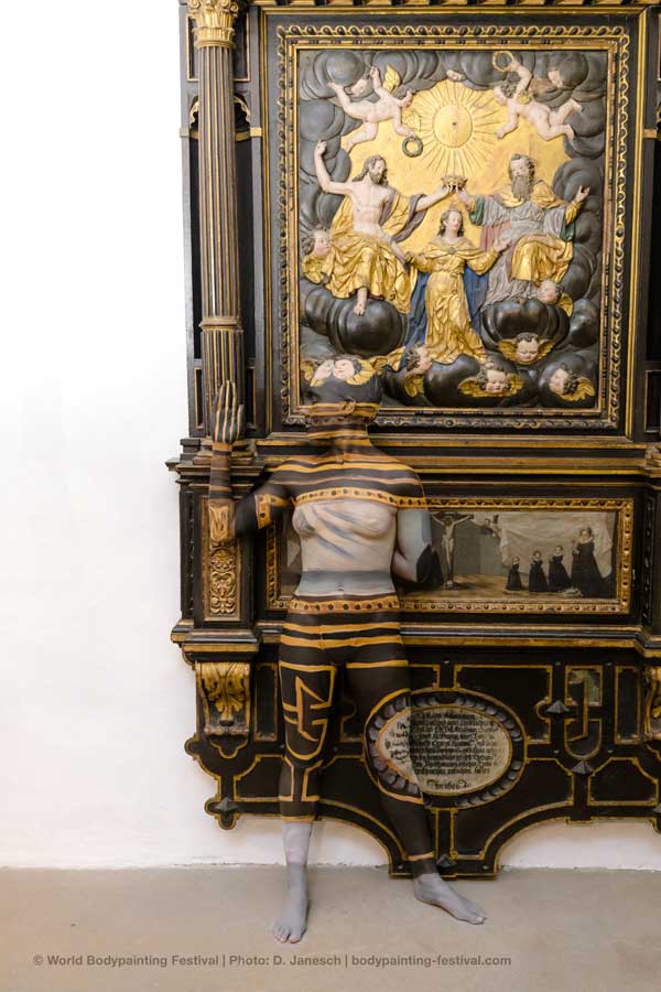 Frau in Leoparden-Bodypainting und großem, mit Früchten geschmücktem Hut posiert am Wasser in der Sonne.