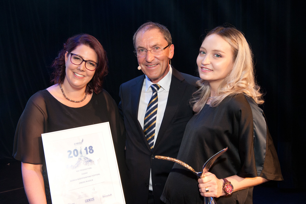 Drei Personen bei einer Preisverleihung. Eine Frau hält eine Urkunde mit der Aufschrift „2018“, eine zweite Frau hält eine goldene Trophäe in Form eines Horns, daneben steht ein Mann im Anzug und gestreifter Krawatte. Alle lächeln in die Kamera.