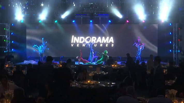Bühne mit der Aufschrift „Indorama Ventures“ im Hintergrund, davor treten bemalte Körperkünstler*innen in leuchtenden Farben auf, während das Publikum an Tischen im Dunkeln sitzt.