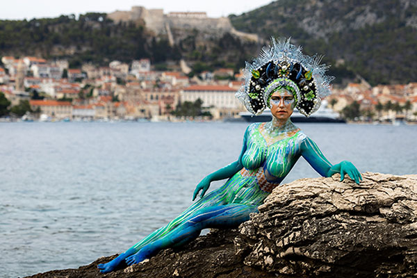 Frau mit kunstvollem Bodypainting in Blau- und Grüntönen, das an Wasser und Natur erinnert, sitzt auf einem Felsen am Meer mit Stadt und Festung im Hintergrund.