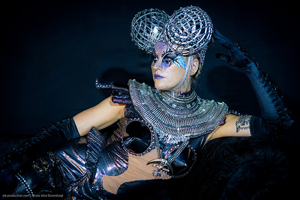 Frau in futuristischem Bodypainting und silbernem Kostüm mit aufwendigem Kopfschmuck aus Metallkugeln, posiert dramatisch auf einem schwarzen Hintergrund.