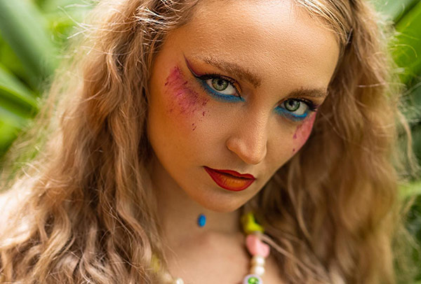 Porträt einer Frau mit auffälligem, buntem Make-up, intensivem Blick und langen blonden Haaren, aufgenommen in tropischer Umgebung.