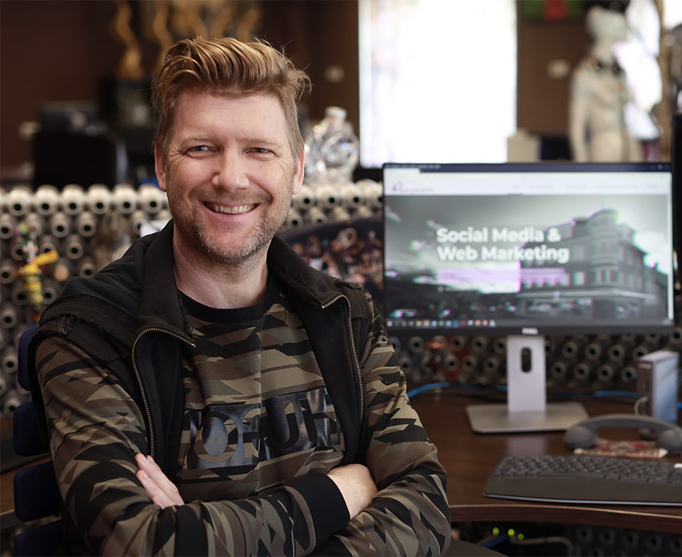 Porträt von Alex Barendregt im Büro, der vor einem Computer sitzt. Auf dem Bildschirm hinter ihm ist die Aufschrift ‚Social Media & Web Marketing‘ zu sehen.