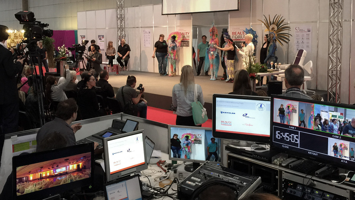 Bühnen- und Backstage-Szene beim Beauty Forum in Leipzig: Modelle mit farbenfrohem Bodypainting stehen auf der Bühne, während Kamerateams und Regie im Vordergrund die Veranstaltung überwachen.