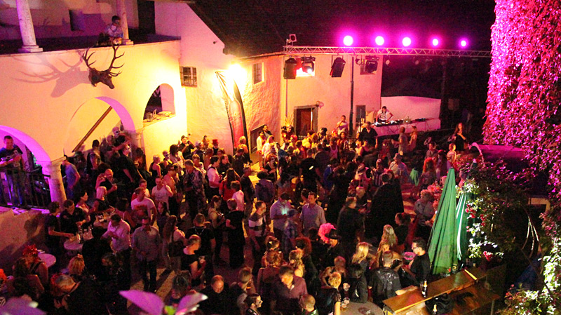 Abendliche Party im Innenhof mit vielen Menschen, DJ-Bühne und bunter Beleuchtung. Gäste tanzen und feiern unter pinken Scheinwerfern.