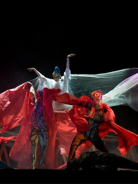 Spektakuläre Bühnenperformance: Drei Tänzer in kunstvollem Bodypainting und Kostümen. Im Vordergrund zwei Figuren mit bunten, tierähnlichen Mustern und roten Tüchern, im Hintergrund eine weiß gekleidete Gestalt mit blauer Kopfbemalung und weit ausgebreiteten Armen.
