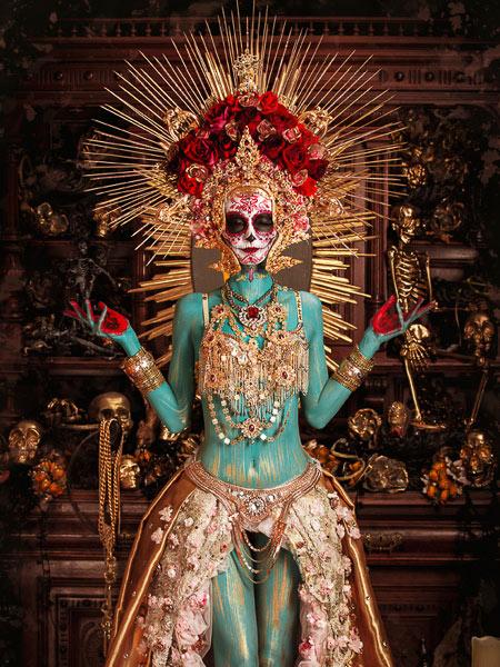 Künstlerisch inszenierte Figur im Stil des ‚Día de los Muertos‘: Eine Person mit türkis bemaltem Körper, goldgeschmücktem Kleid und aufwendig gestaltetem Totenkopf-Make-up. Auf dem Kopf trägt sie eine Krone aus roten Rosen und goldenen Strahlen, im Hintergrund sind Schädel und barocke Dekorationen zu sehen.