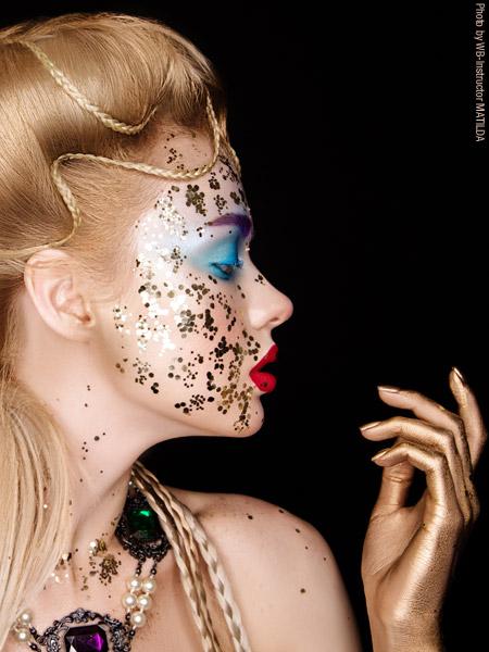 Porträt einer Frau mit aufwendig gestaltetem Make-up und Bodypainting: Ihr Gesicht ist mit goldenem Glitzer und floralen Mustern verziert, die Lippen sind leuchtend rot, die Augenpartie in kräftigem Blau betont. Die Hand ist gold bemalt, Schmuck und geflochtenes Haar runden den glamourösen Look ab