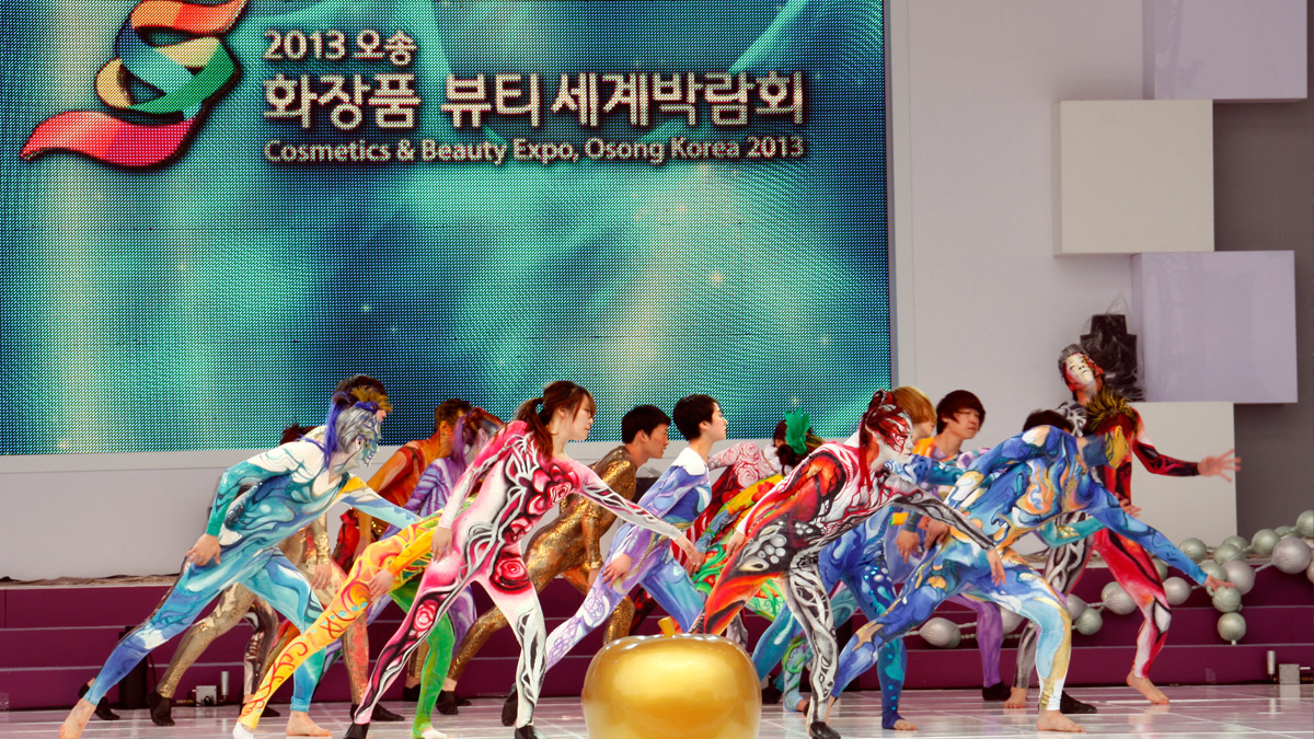 Tanzperformance bei der Cosmetics & Beauty Expo 2013 in Osong, Korea: Eine große Gruppe von Tänzern in farbenfrohem Bodypainting bewegt sich synchron vor einer LED-Bühnenwand.