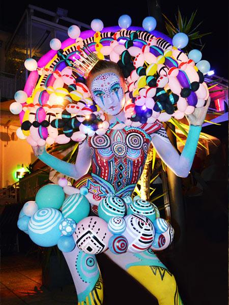 Farbintensive Bodypainting-Performance: Eine Person ist in kräftigen Mustern und Neonfarben bemalt und trägt eine spektakuläre Kostümkonstruktion aus bunten Ballonformen, die wie ein leuchtender, ornamentaler Kopfschmuck wirkt.