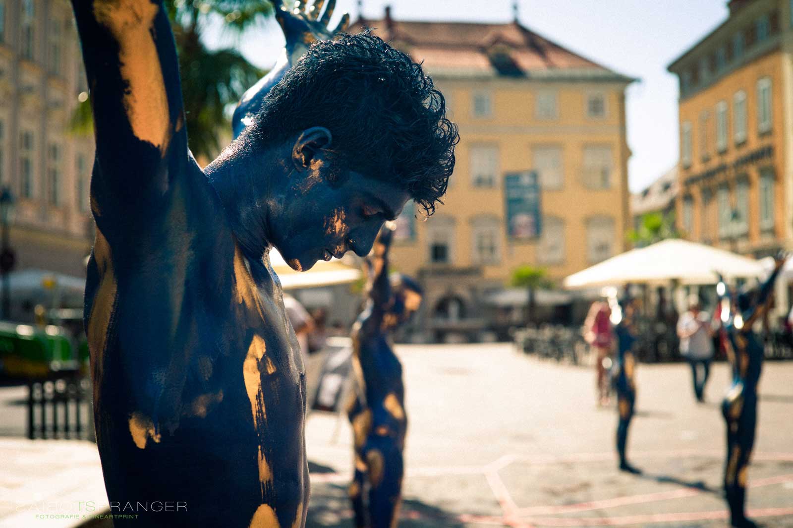 Körperbemalter Mann mit blau-goldener Farbe steht mit gesenktem Kopf und erhobenen Armen im Freien. Im Hintergrund weitere bemalte Personen und eine historische Stadtarchitektur.
