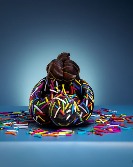 Körperbemalung in Form eines Donuts mit bunter Zuckerglasur und Streuseln, die wie ein echtes Gebäckstück wirkt, umgeben von verstreuten Streuseln auf blauem Untergrund.
