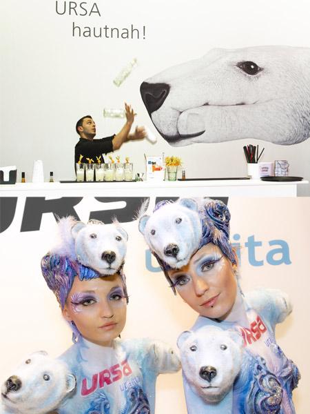 Messestand mit Eisbär-Motiv: Oben jongliert ein Barkeeper vor einer Wandgrafik mit großem Eisbärkopf und dem Schriftzug ‚URSA hautnah!‘. Unten stehen zwei Frauen in Bodypainting-Kostümen mit Eisbärköpfen und blauer Bemalung, passend zum Markenauftritt.