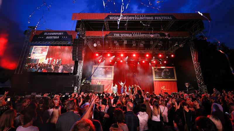 Bühnenaufnahme vom World Bodypainting Festival in Klagenfurt: Eine große Menge feiert vor der rot beleuchteten Hauptbühne mit Leinwänden und Performances.