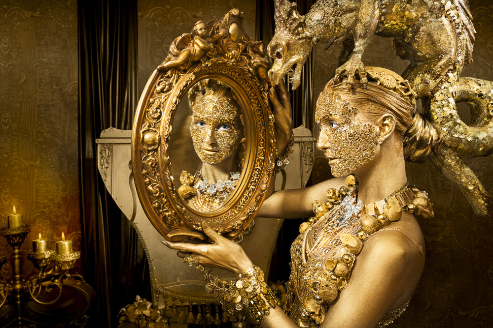 Frau in goldener Körperbemalung mit funkelnden Schmuckelementen hält einen prunkvollen, goldenen Spiegel, in dem ihr Gesicht reflektiert wird. Über ihr ragt eine goldene Drachenfigur, während Kerzenständer und barocke Dekoration den opulenten Hintergrund bilden.