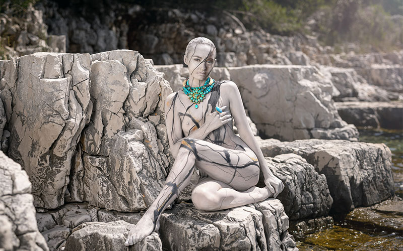 Menschliche Figur, kunstvoll bemalt und posiert wie ein Schokoladendonut mit bunten Zuckerstreuseln, vor blauem Hintergrund.